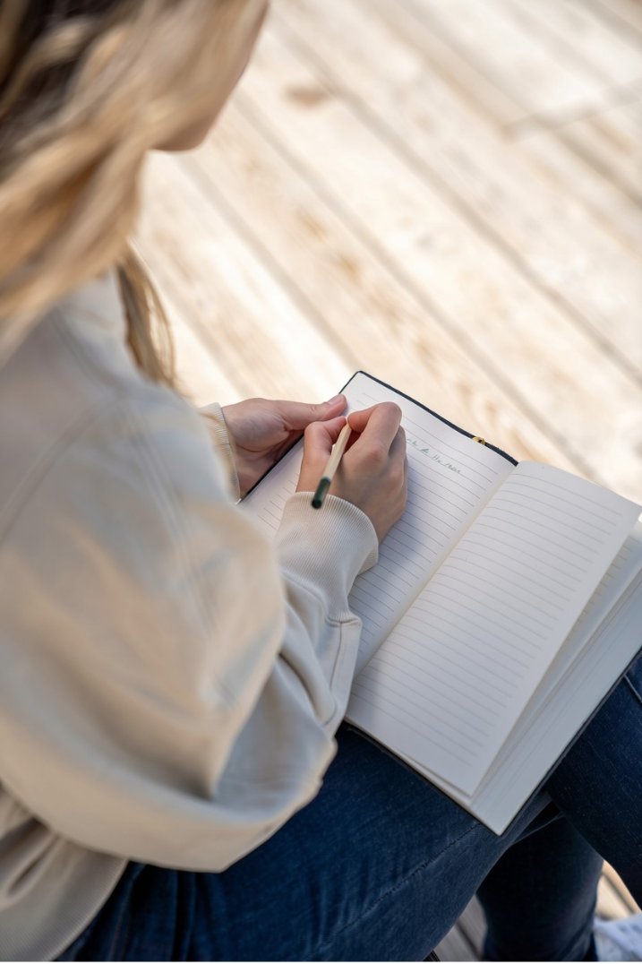 Osoba widziana zza ramienia zapisuje coś w notesie opartym na kolanach