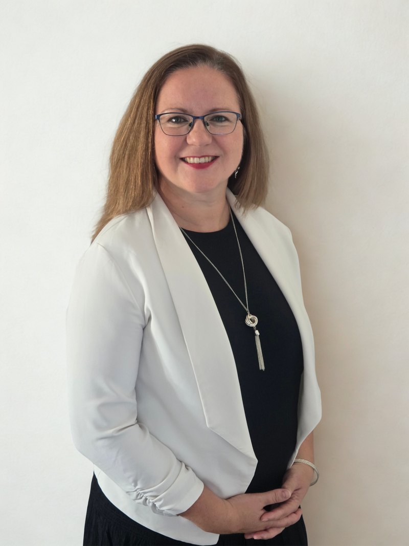 Joanna Laszczak w białej marynarce, uśmiechnięta, z dłońmi złożonymi przed sobą